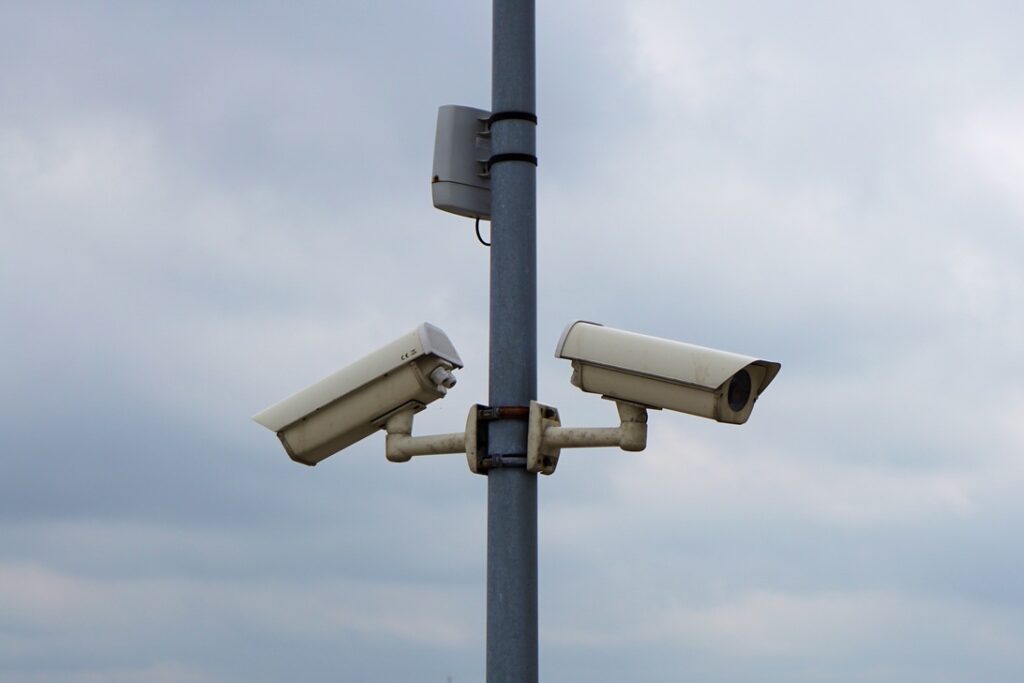 Two white security cameras mounted on a metal pole against a cloudy sky, monitoring an outdoor area.
