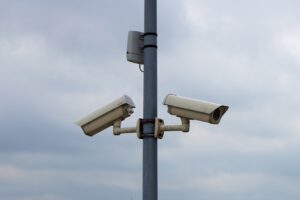 Two white security cameras mounted on a metal pole against a cloudy sky, monitoring an outdoor area.