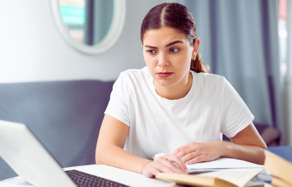 Woman in a white t-shirt sits at a desk with a laptop and papers, looking thoughtful.