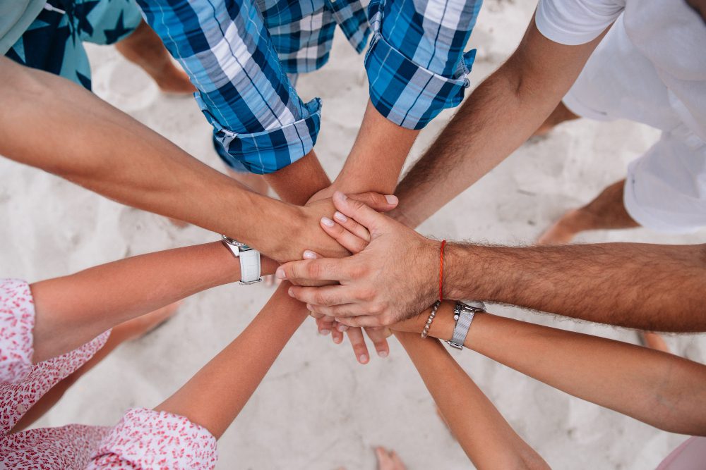 Group of diverse hands stacked together in a circle on a sandy beach, symbolizing teamwork and unity.