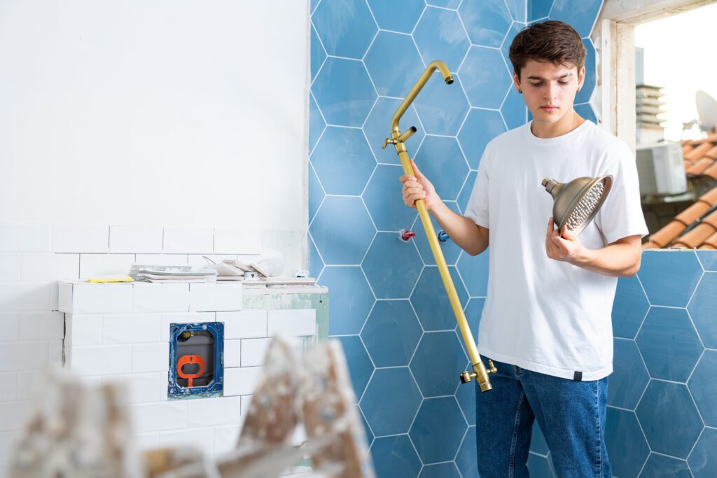 Young man in a white shirt and jeans installs a brass shower arm in a bathroom with blue hex tile walls (renovation scene).