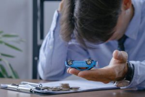 Person in business attire holds a small blue toy car in their palm over a desk with coins and documents, illustrating auto budgeting or insurance planning.