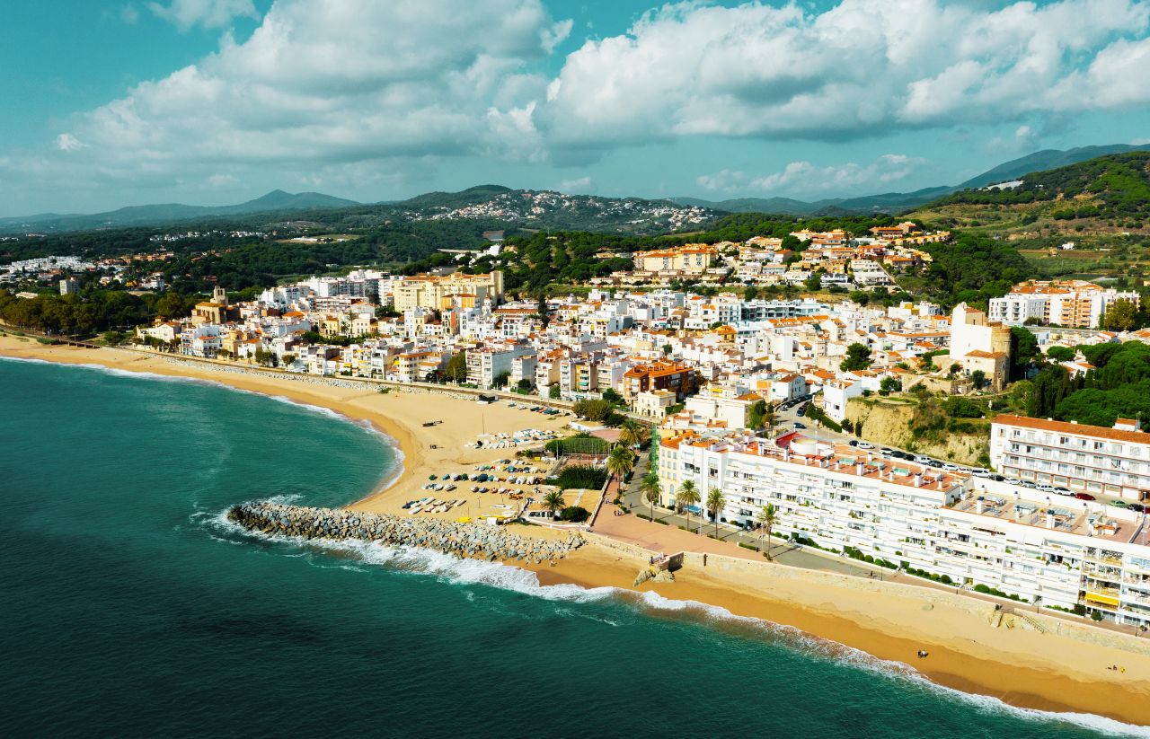 763809 - Finques Sant Pol - Barcelona Noticias 24h Coastal town with white buildings along a sandy beach and turquoise sea backed by green hills in the distance.
