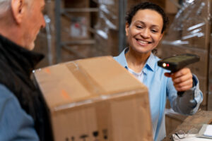 Warehouse worker scans a cardboard box with a handheld scanner while a man hands it to her, smiling in a warehouse setting.