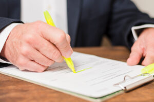 Close-up of a hand in a suit signing a document with a bright yellow highlighter on a clipboard on a wooden desk.