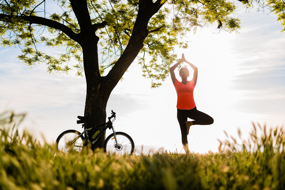 Person doing a yoga pose (tree pose) beside a bicycle leaning against a large tree in a sunlit park field.