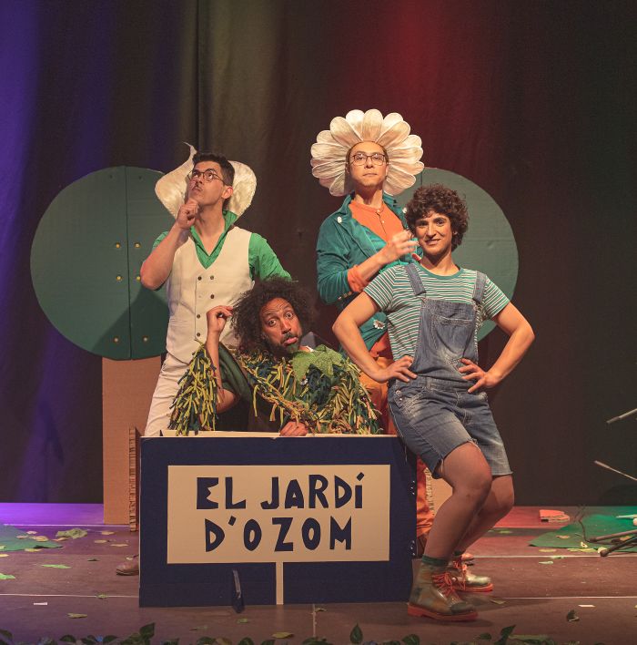 A group of four performers on a stage in colorful costumes, with a sign reading 'EL JARDI D' OZOM' in front, posing playfully as a garden scene unfolds.