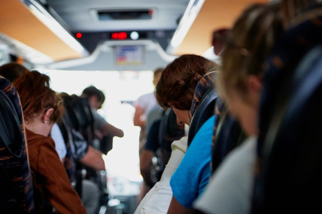 People seated in a bus aisle, some looking down or away, sunlit doorway at the far end behind them.