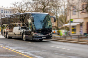 Black tour bus driving down a city street, its reflective surface showing surrounding buildings and trees in the glare of the day.