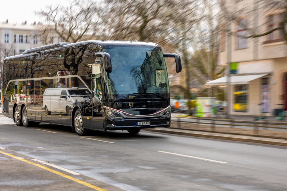 Black tour bus driving down a city street, its reflective surface showing surrounding buildings and trees in the glare of the day.
